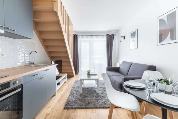 A modern kitchen with grey cabinets, a wooden staircase, a dining area with a grey rug, and a grey sofa.