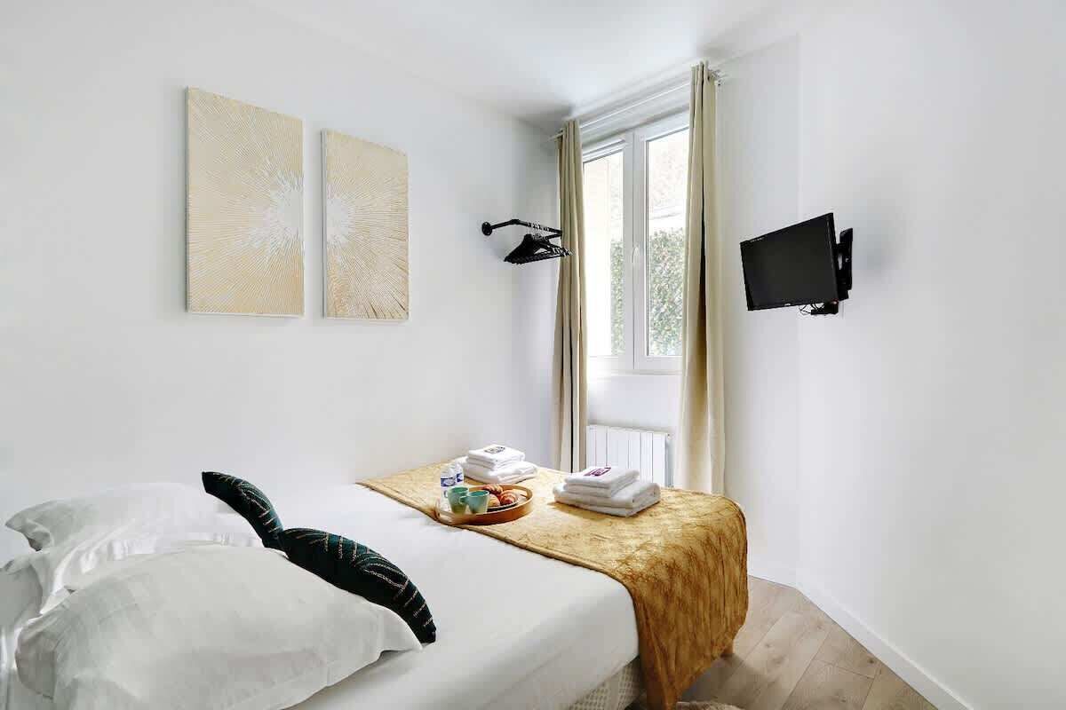 A hotel room with a bed, a small table with towels and a cup, a wall-mounted TV, and two abstract paintings.