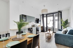 A modern living room with a blue sofa, dining area with a table set for two, and a kitchen area with appliances. - Appartement de charme proche de la rue Lecourbe (Paris)