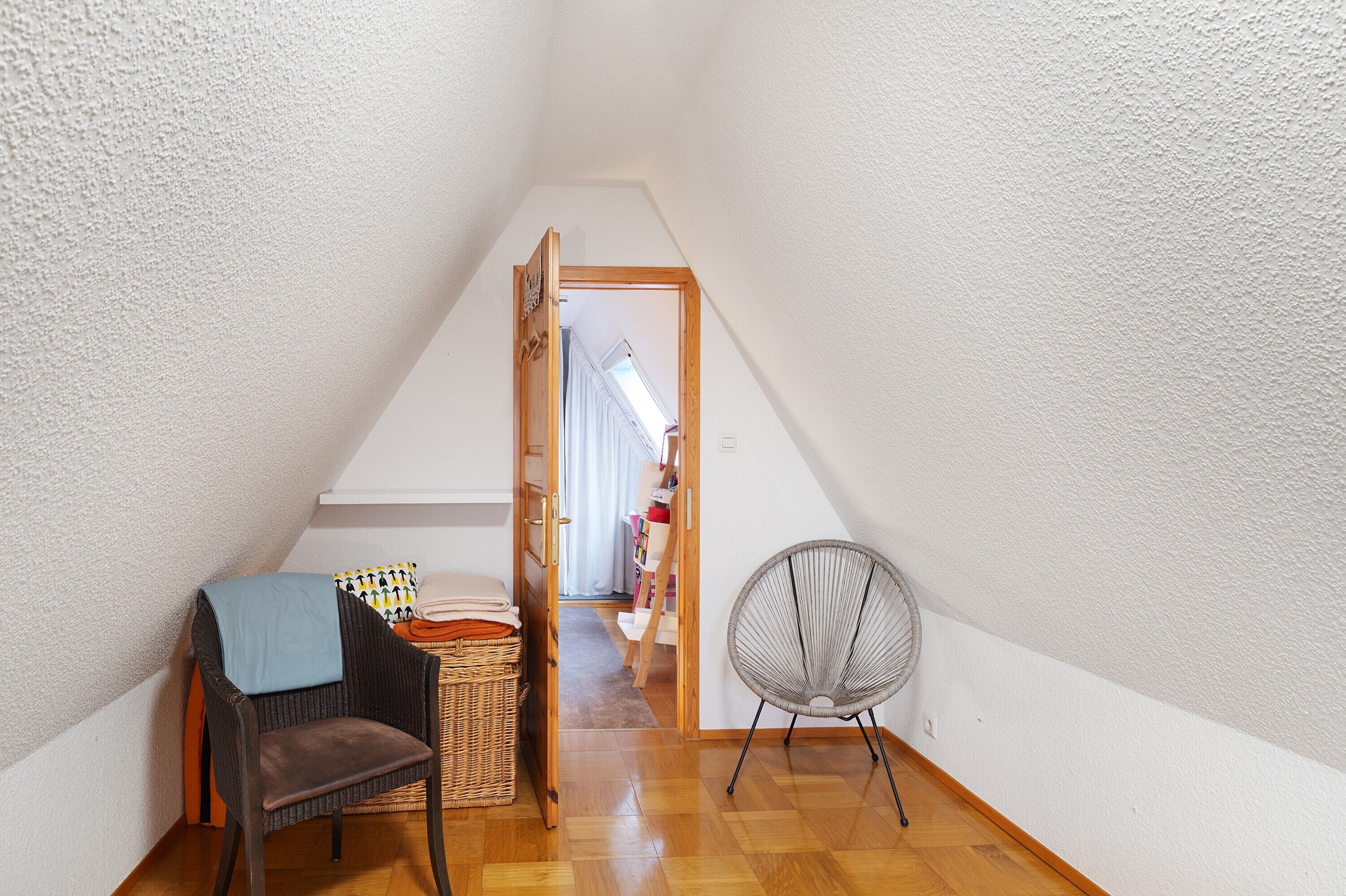 A narrow, sloped room with a wooden floor, a wicker chair, a fan, and a chair with a blanket.