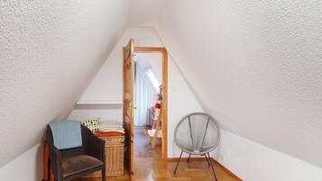 A narrow, sloped room with a wooden floor, a wicker chair, a fan, and a chair with a blanket.