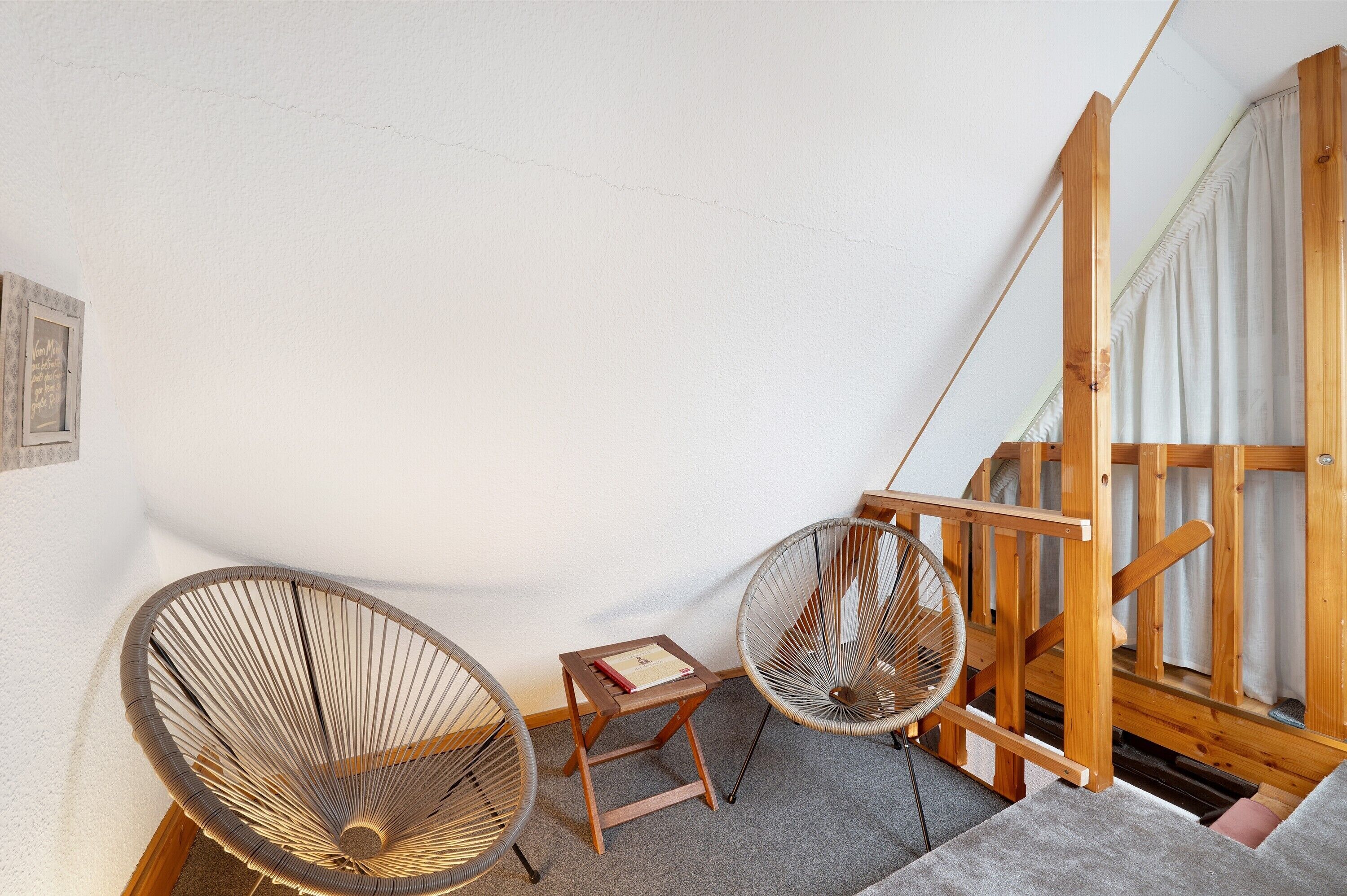 Une pièce avec un escalier en bois, deux fauteuils en osier, une petite table en bois et un mur blanc.