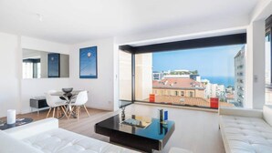 A modern living room with a dining area, large windows with a city view, and a balcony. - Appartement de Luxe 3BR vue  sur la Mer (Beausoleil)