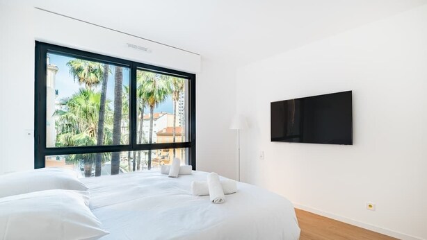 A hotel room with a large window, a bed with white linens, a flat-screen TV mounted on the wall, and a wooden floor. - Appartement de Luxe 3BR vue  sur la Mer (Beausoleil)