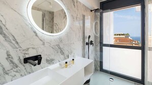 A modern bathroom with a marble backsplash, a large mirror, a shower area, and a window with a view of buildings and the sea. - Appartement de Luxe 3BR vue  sur la Mer (Beausoleil)