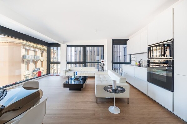 Apartamento moderno con sala de estar, cocina y ventanas amplias que ofrecen vistas de un edificio.