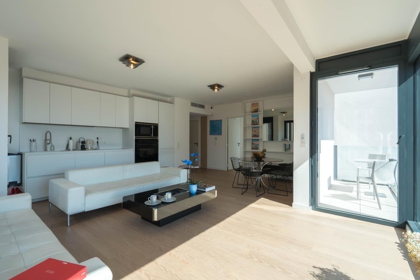 Een moderne leefruimte met een witte bank, een eethoek met tafel en stoelen, en een keuken met kasten en apparatuur.