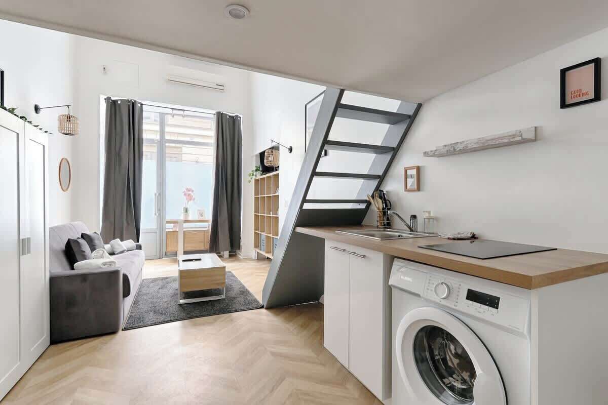 A compact living space with a sofa, a washing machine, a kitchen area, and a staircase leading to an outdoor balcony.
