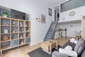 Una sala moderna con sofá, mesa de centro y estantería. Se ve una escalera que sube a un nivel superior con cocina y lavadora.