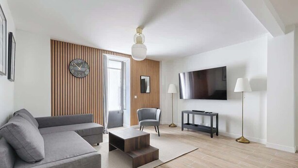 A modern living room with a grey sofa, a coffee table, a TV, and a wooden accent wall. - Batignolles Epinette Lovely Apartment 6P (Paris)
