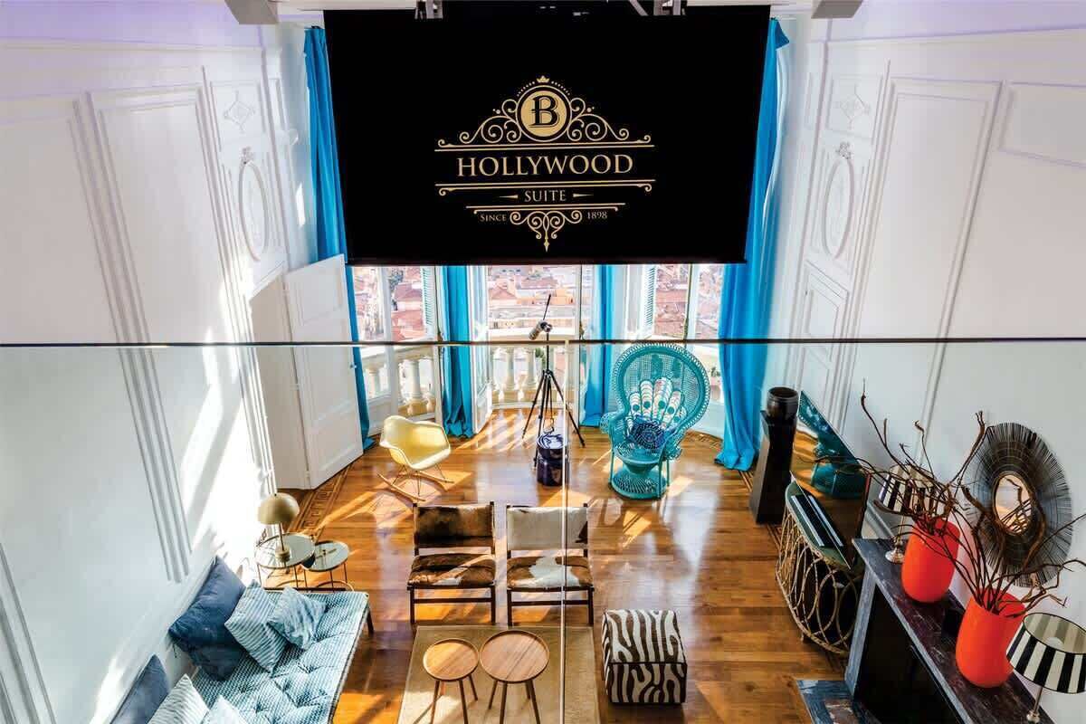Un suite d'hotel amb una finestra gran, un conjunt de sofàs, una taula de dinar i una pantalla de televisió que mostra el nom de l'hotel.