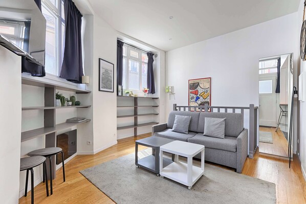 A modern living room with a grey sofa, a white coffee table, and a grey rug. There are shelves with plants and a window with curtains.