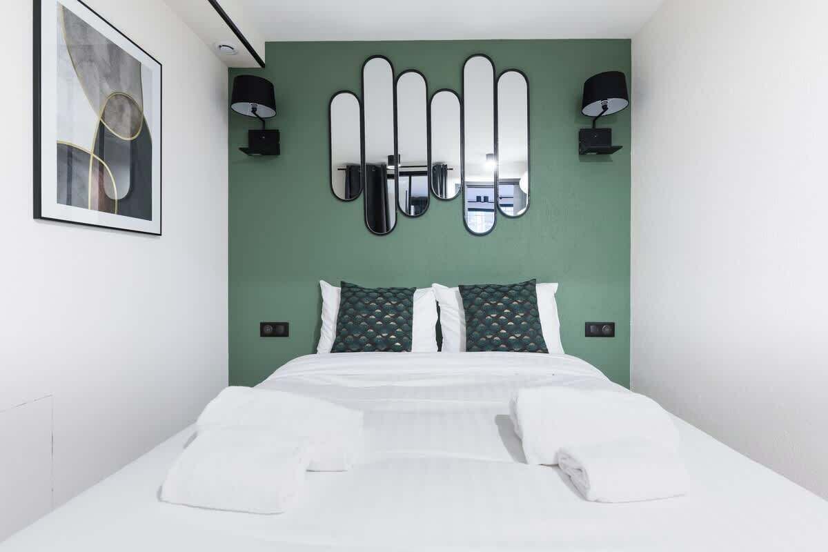 A hotel room with a bed, green walls, a framed abstract artwork, and a wall-mounted mirror.