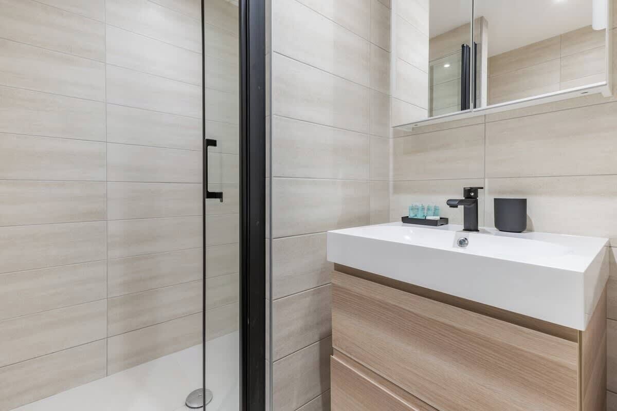 Une salle de bain moderne avec une douche à l’italienne en verre, un lavabo blanc avec un robinet noir et un miroir au-dessus du lavabo.