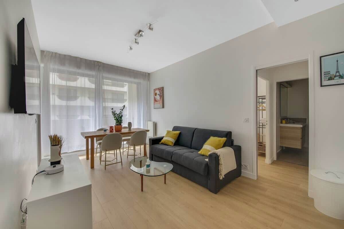 Ein modernes Wohnzimmer mit einer Couch, einem Couchtisch und einem Essbereich. Ein Fernseher ist an der Wand montiert und es hängt ein Gemälde vom Eiffelturm.