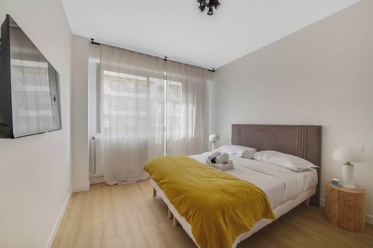 A modern bedroom with a bed, a bedside lamp, and a window with sheer curtains.