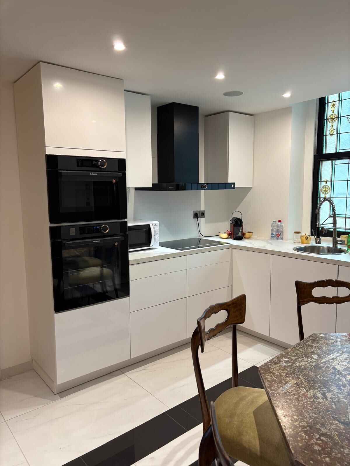 A modern kitchen with white cabinetry, a black oven, a microwave, and a stainless steel sink. There is a dining table with chairs and a window with decorative glass.