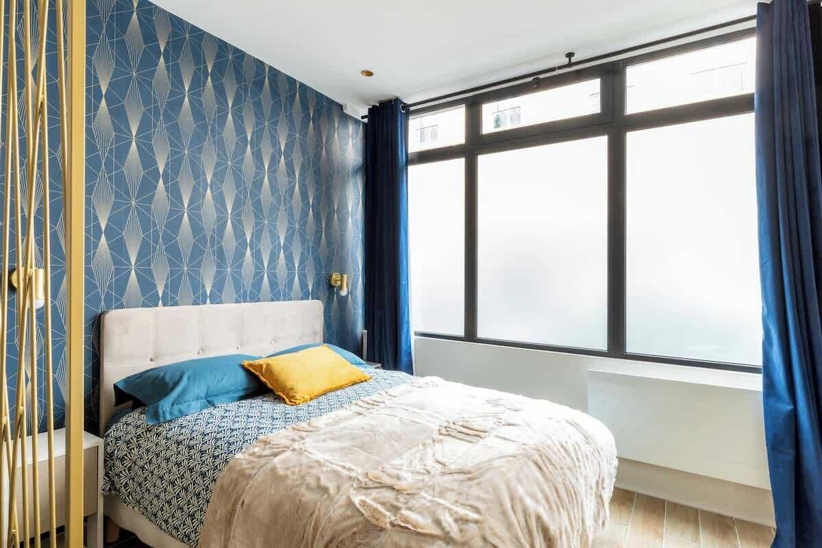 A bedroom with a geometric patterned wall, a large window with curtains, and a bed with patterned bedding.