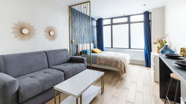 Un soggiorno moderno con un divano grigio, un tavolino da caffè bianco e un letto con testiera blu. Si nota un'ampia finestra con tende azzurre, un elemento decorativo a parete e un piccolo angolo cottura con uno sgabello da bar.