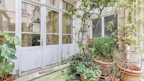 Un patio con plantas en macetas, una puerta blanca y un ventanal con vista a un jardín.
