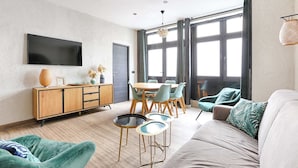 Ein modernes Wohnzimmer mit Flachbildfernseher, einem Essbereich mit Holztisch und Stühlen sowie einem großen Fenster mit Vorhängen.