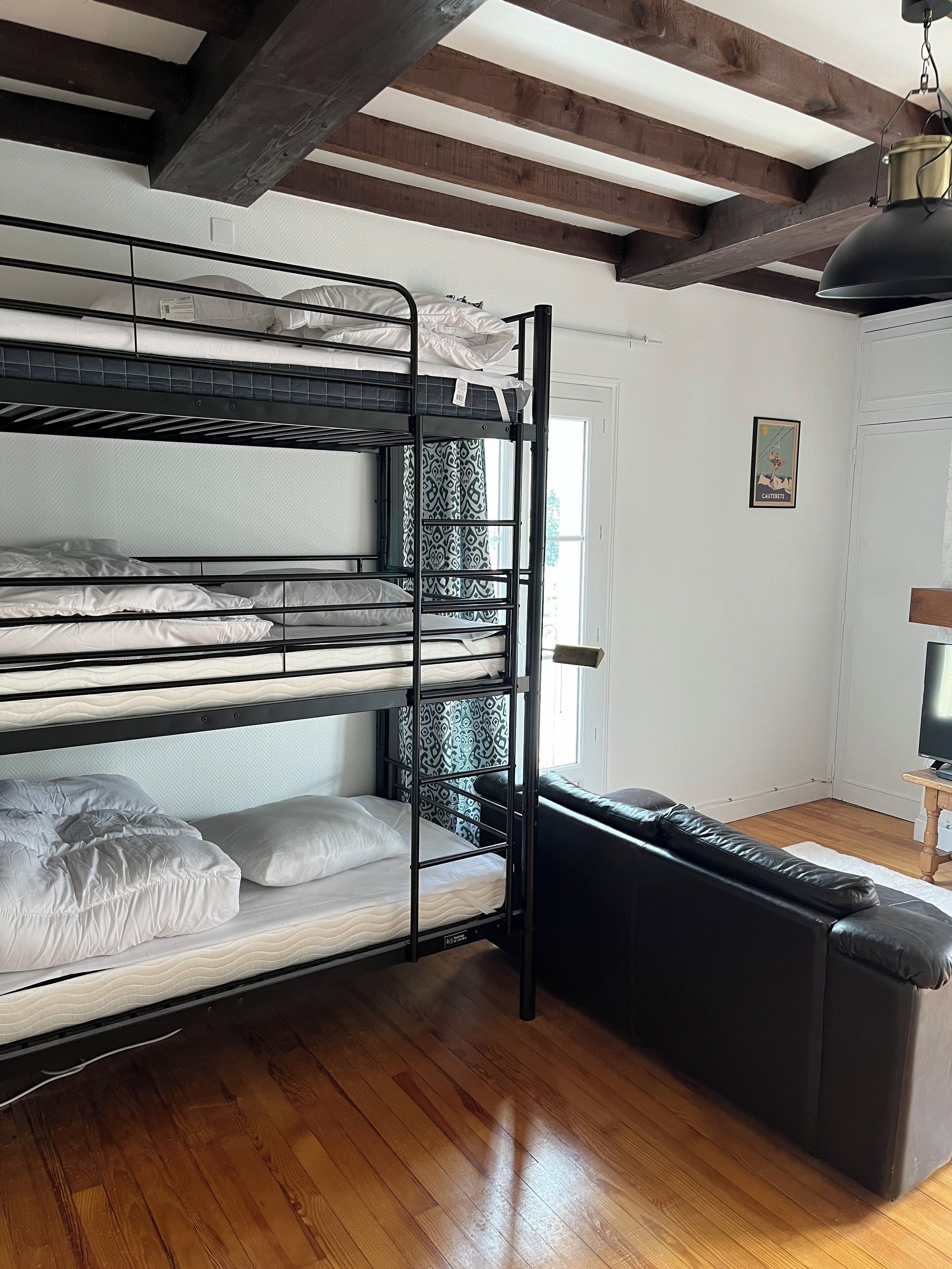 Una cama tipo litera con tres niveles, un sofá de cuero negro, suelo de madera y una puerta blanca.