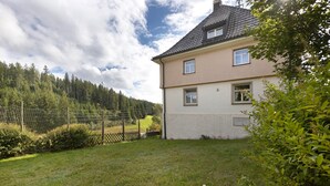 Ein zweistöckiges Haus mit geneigtem Dach, einem eingezäunten Garten und einem bewaldeten Hügel im Hintergrund.