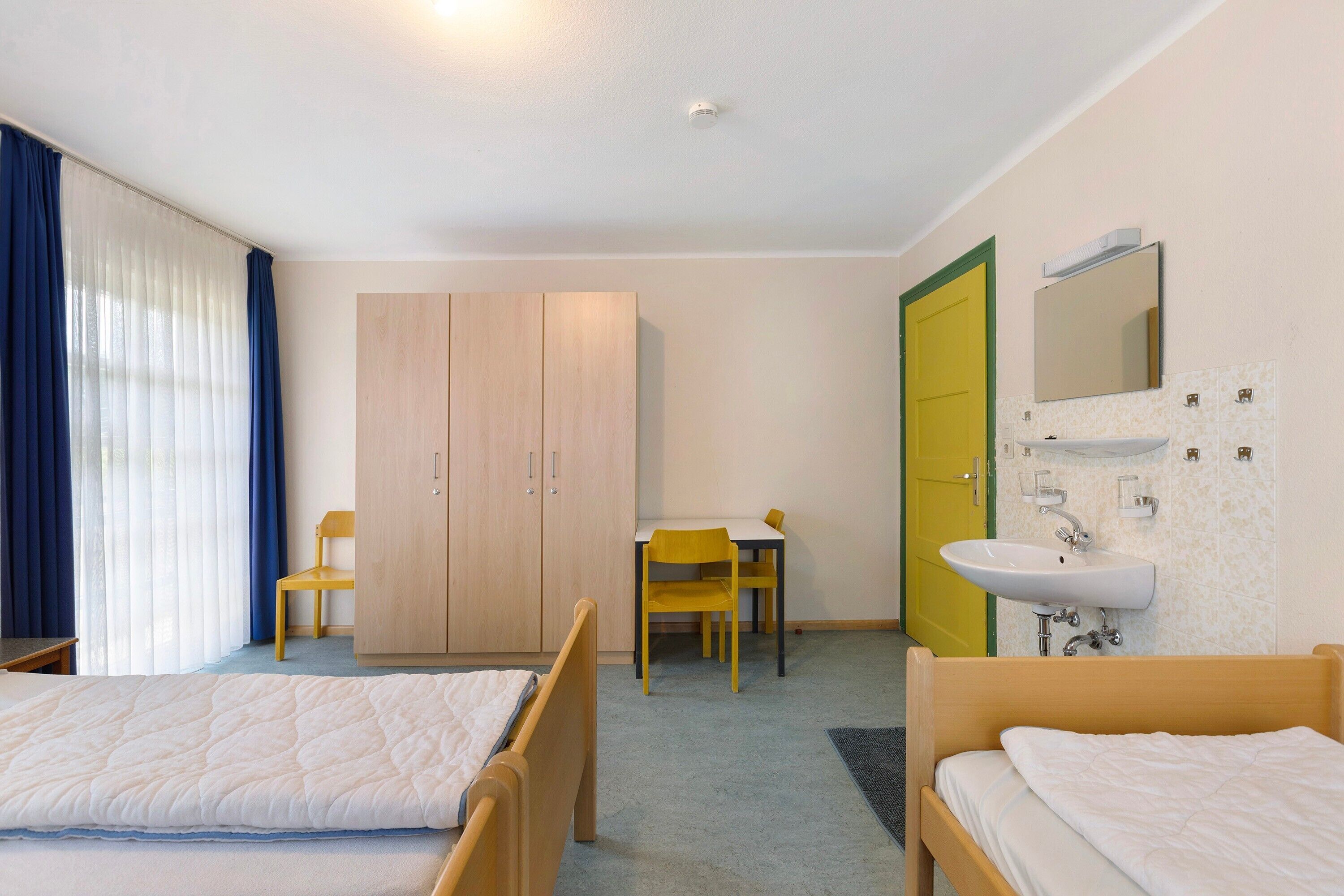 Une chambre d’hôtel avec deux lits, un bureau, une chaise, une armoire, une salle de bain avec un lavabo et un miroir, et une porte donnant sur une autre pièce.