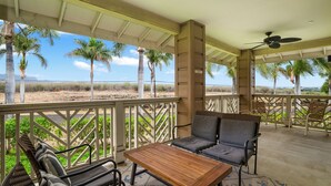 Un espacio de estar al aire libre techado, con una mesa y sillas de madera, un ventilador de techo y vistas a la playa y palmeras.