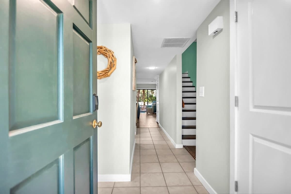 Un couloir avec une porte, une couronne décorative accrochée au mur, un escalier et une vue sur une pièce.