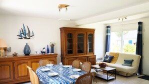 Un coin repas avec une table ronde dressée pour quatre personnes, entourée de chaises en bois. Un buffet en bois avec des portes en verre se trouve à gauche, et un canapé avec un coussin est placé à droite.