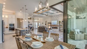 Une cuisine moderne avec des meubles blancs, un coin repas doté d’une table en bois pour quatre personnes et un salon avec des meubles en osier.