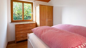 A bedroom with a wooden wardrobe, a bed with a checkered bedspread, a window with a view of greenery, and a wooden ceiling.