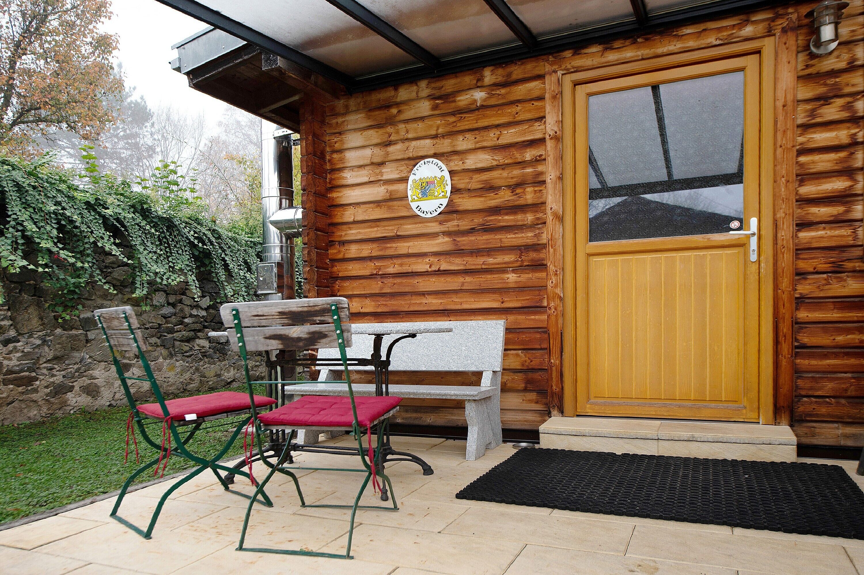 A wooden exterior with a door, a bench, and two chairs with red cushions.