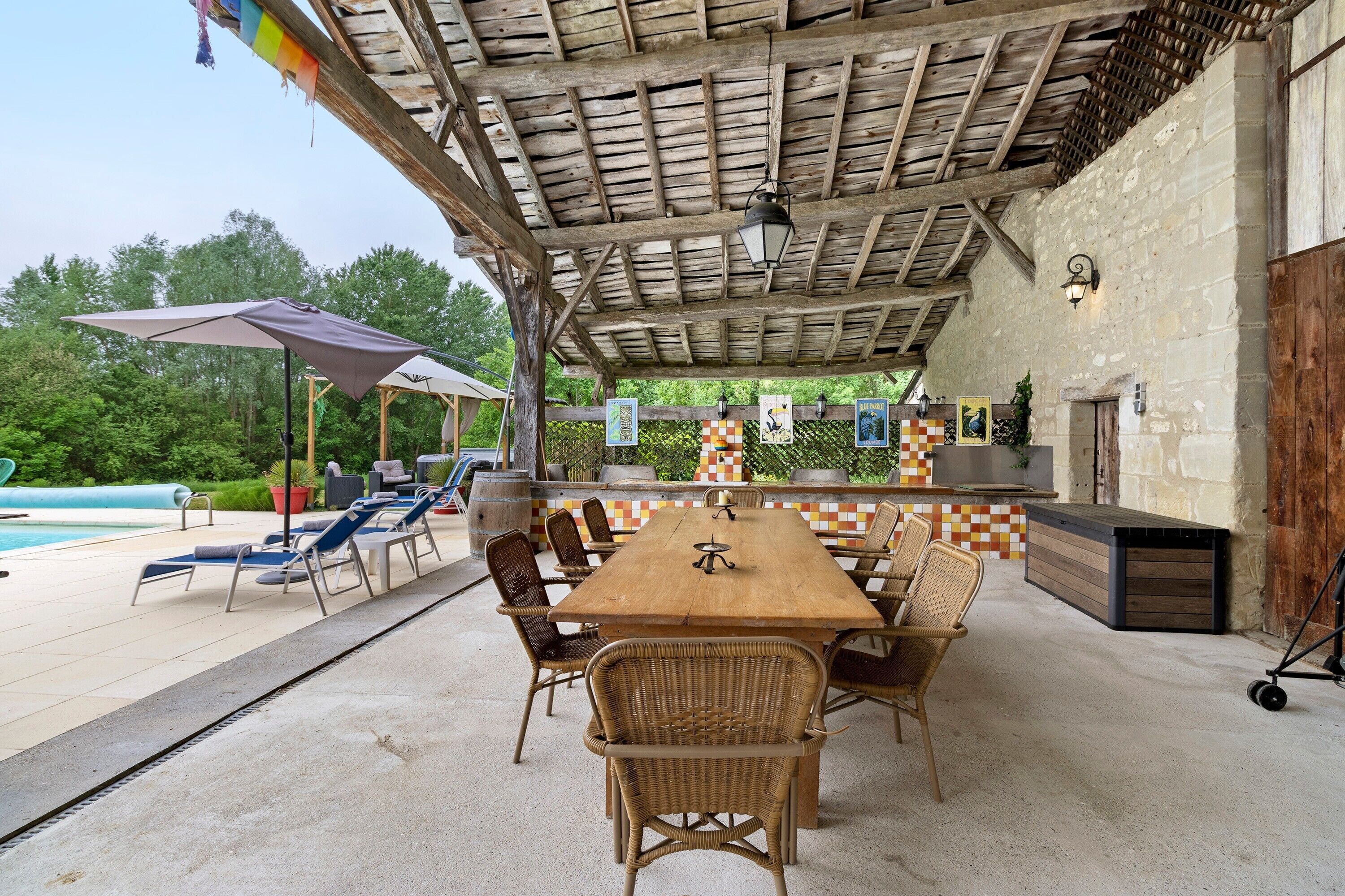 Une terrasse extérieure avec une table en bois et des chaises en osier, une piscine et un espace barbecue.