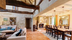 A spacious interior with a high ceiling, exposed wooden beams, a dining area with a long table and chairs, and a fireplace.