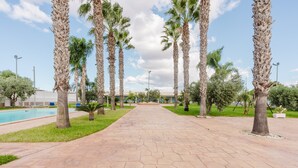 A paved pathway lined with palm trees, a swimming pool, and a building in the background. - Holiday Home 'La Perla Di Mare - Giada' with Shared Pool, Wi-Fi and Air Conditioning (Mazara Del Vallo)