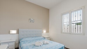 A bedroom with a bed, bedside tables, a nightstand, a wall-mounted light, and a window with bars. - Holiday Home 'La Perla Di Mare - Giada' with Shared Pool, Wi-Fi and Air Conditioning (Mazara Del Vallo)
