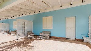 Una habitación con pared azul, puerta blanca y ventana con contraventanas. Hay sillones de mimbre y una mesa.