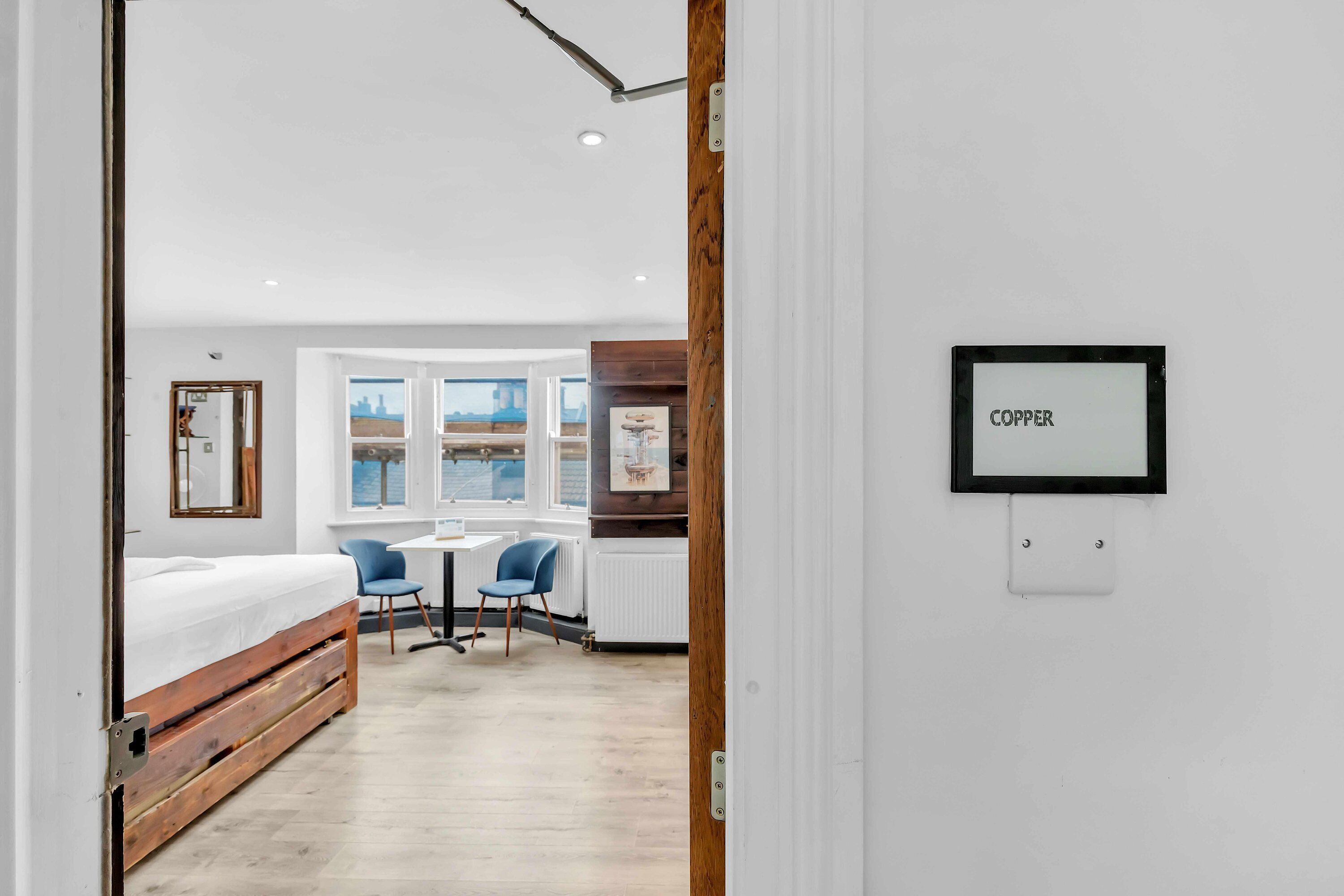 A modern room with a bed, a desk, and a chair. There is a window with a view of the city.