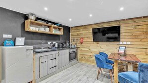 A modern kitchen with grey cabinets, a wooden countertop, and a built-in oven. There is a wooden wall with a mounted TV and a small table with blue chairs. - Grafton Woodshack | By My Getaways (Brighton)