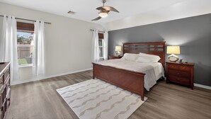 A bedroom with a wooden bed, two bedside tables with lamps, a ceiling fan, and a view of the outdoors through a window with white curtains.