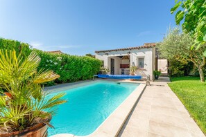 A swimming pool with a tiled deck, surrounded by greenery and a building with a tiled roof.