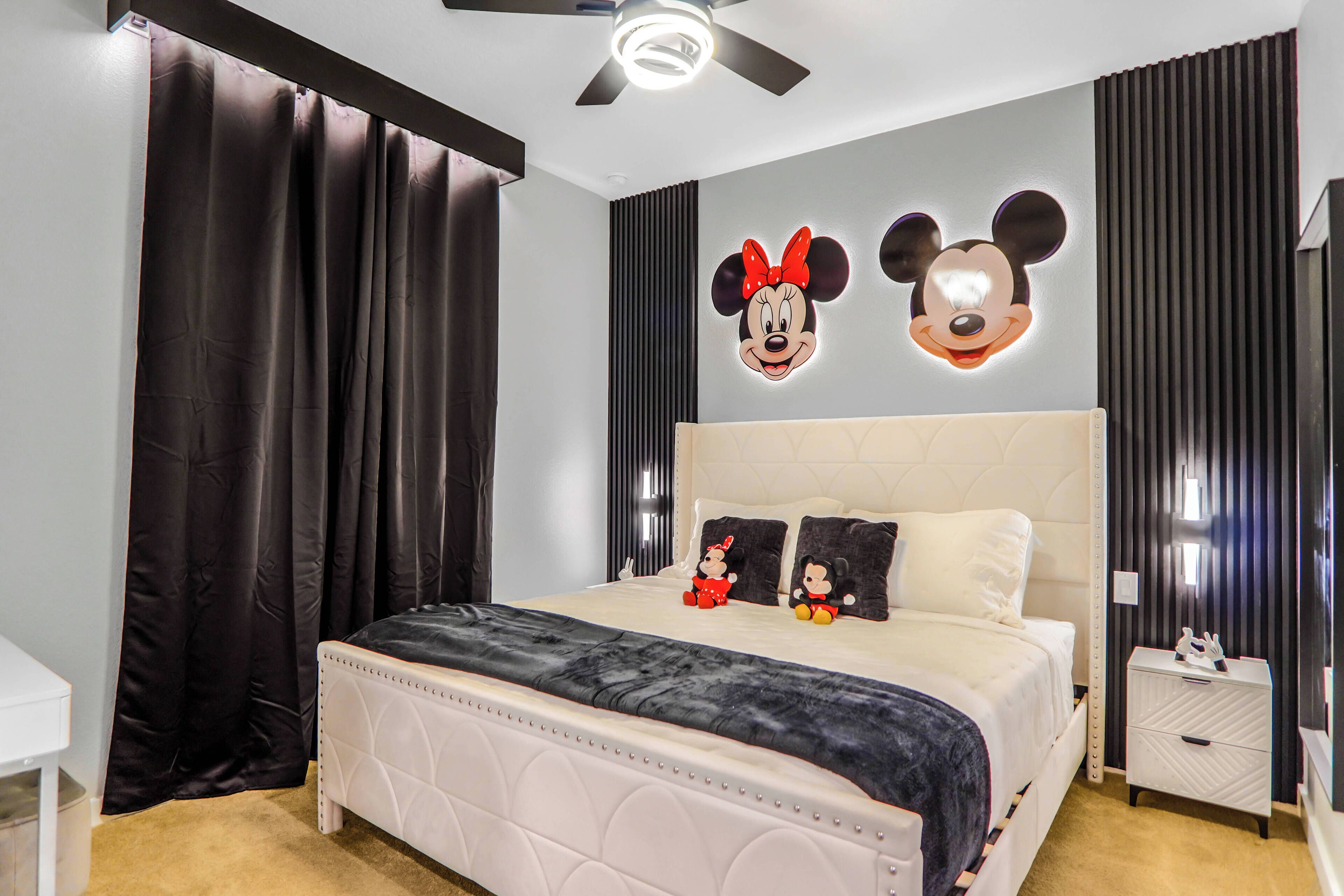 Une chambre à coucher avec un grand lit, des rideaux foncés, un ventilateur de plafond et deux décorations murales représentant Mickey Mouse, le tout éclairé.