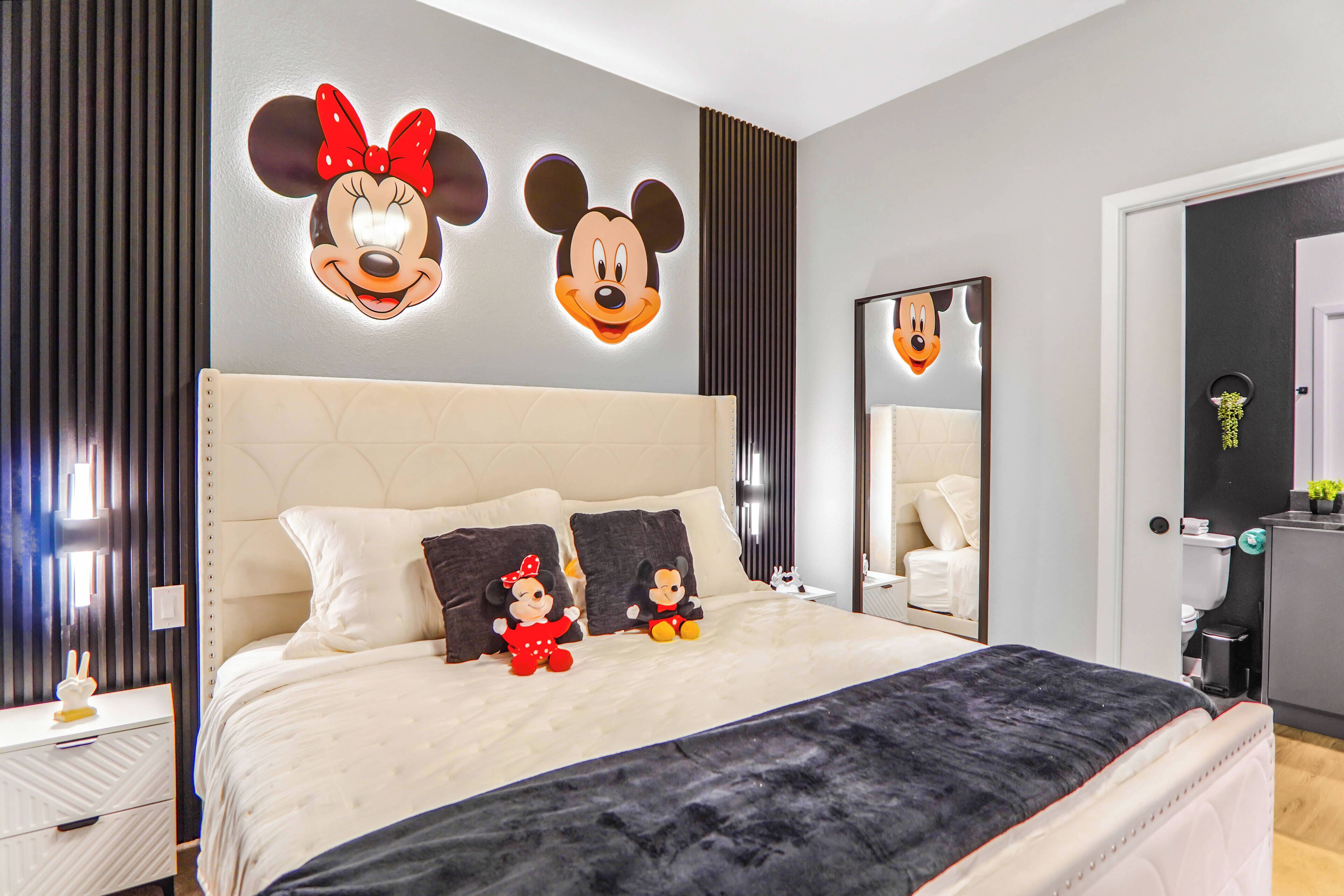 A hotel room with a large bed, two Mickey Mouse wall decorations, a mirror, and a bedside table with a lamp.