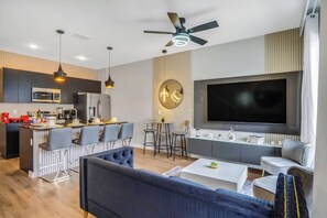 A modern kitchen with a microwave, refrigerator, and dining area. There is a large flat-screen TV mounted on the wall, a ceiling fan, and a bar area with stools.