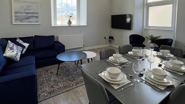 Un salon moderne avec un canapé bleu foncé, une table à manger dressée pour six personnes et de grandes fenêtres en arc de cercle.