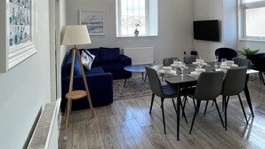 Un salon moderne avec un canapé bleu, une table à manger dressée pour six personnes et une grande fenêtre en arc de cercle.