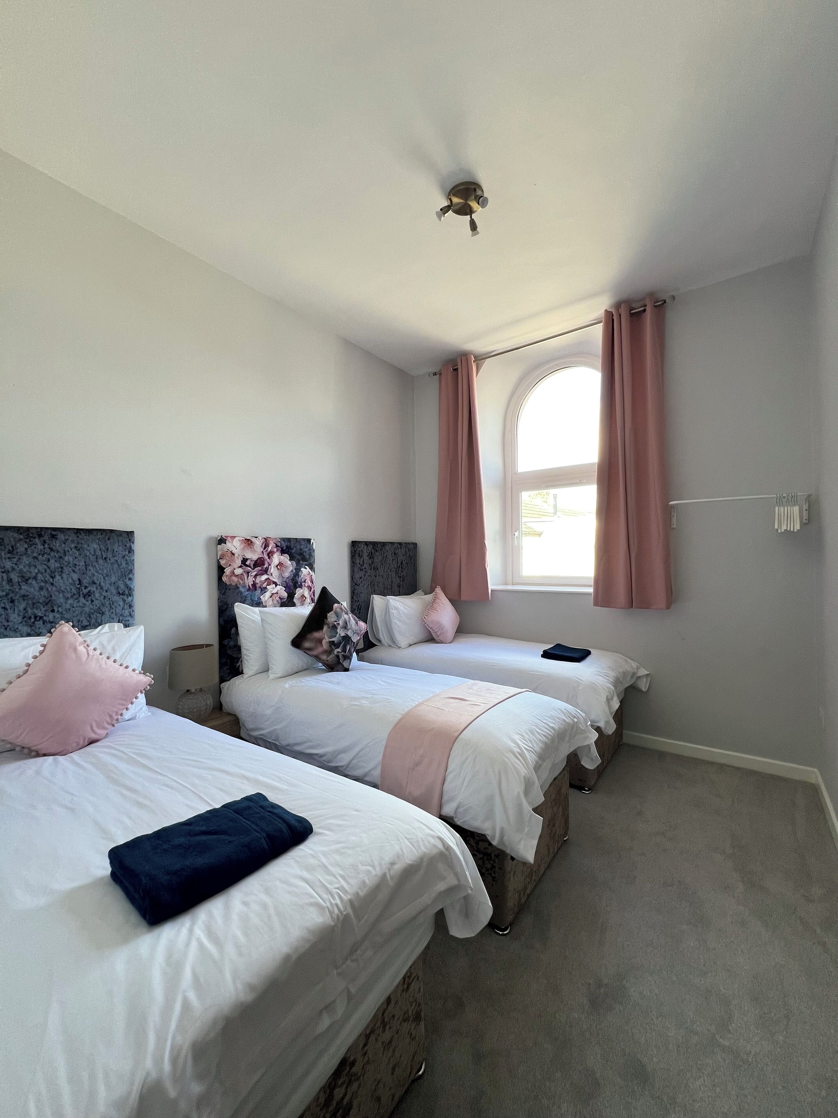 Quarto com duas camas, ventilador de teto, janela com cortinas e mesa de cabeceira com abajur.