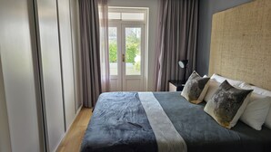 Une chambre à coucher avec un grand lit, des rideaux gris, une table de chevet avec une lampe et une vue sur l’extérieur, visible à travers une porte.